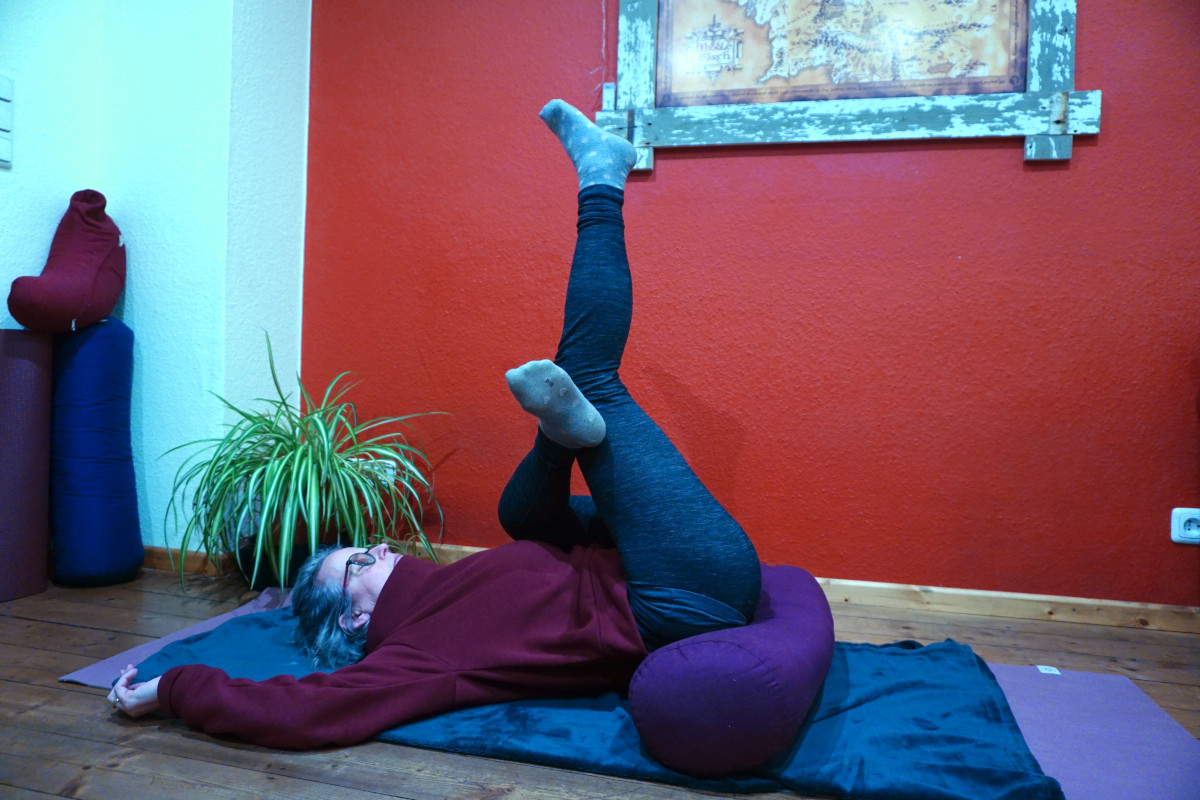 Yin Yoga Position Nadelöhr. Sie gehört zu meinem morgendlichen Ritual. Das Bild zeigt Susanne auf dem Rücken liegend. Das Bolster liegt unter dem Gesäß. Das linke Bein ist angewinkelt vor dem Rechten. Beide Beine fallen durch die Schwerkraft Richtung Oberkörper. Dabei das rechte Beine gestreckt.