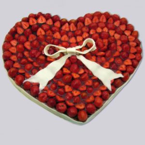 Das Foto zeigt einen Erdbeekuchen in Herzform. Darauf ist eine Schleife aus Teig gelegt.