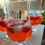 Das Foto zeigt 3 Gläser mit Erdbeercocktail. Der Cocktail ist rötlich und durchscheinend. Oben auf schwimmen einige geviertelte Erdbeeren, Eiswürfel und Minze. Im Hintergrund sind Messer in einem Messerblock zu erkennen.