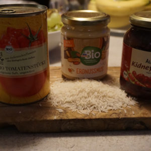 Das Foto zeigt eine Glas Kidneybohnen, Erdnussmuss und eine Dose Tomaten auf einem Holzbrett. Davor ist Reis verstreut.